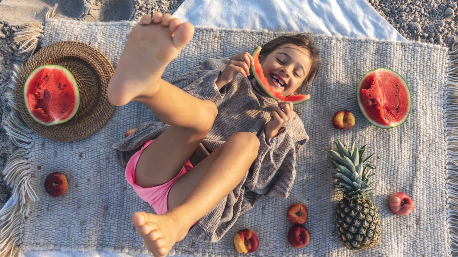 A little girl laughing, while eating a large slice of watermelon on the beach. She is lying on a beach towel with fruit scattered around her.