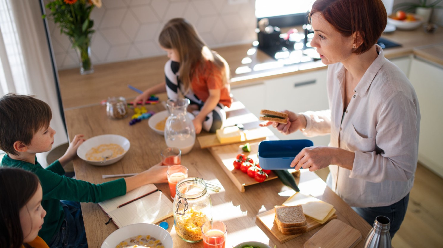 A mother is making school lunch while her three children are eating breakfast at the kitchen bench. It is a busy scene.