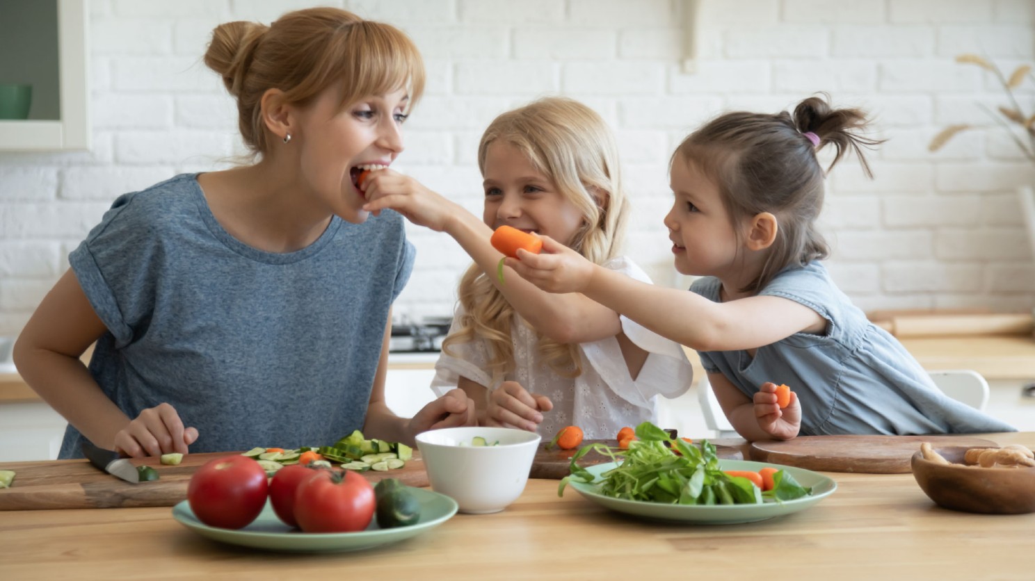 A woman sits with two young children, who are feeding her carrot sticks. They sit at a kitchen bench with plates and bowls full of vegetables. All have smiles on their faces.