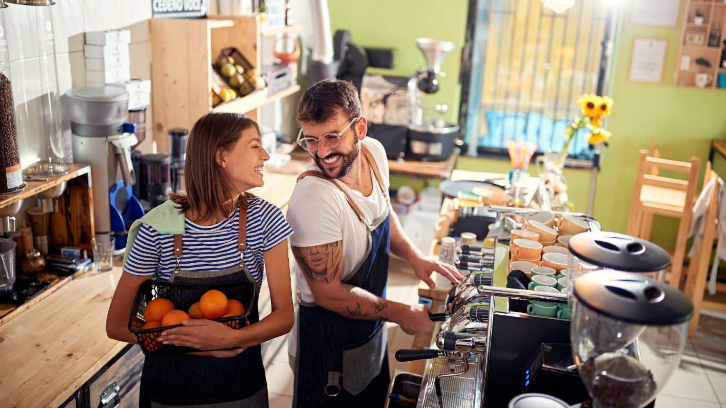 A young man and woman working in a cafe, presumable the cafe owners. The man is making coffee, and the woman is carrying a basket of oranges. They are looking at each other with large smiles on their faces.